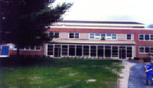 maria (1) A two-story brick building with many windows, a tree on the left, and a person standing on a paved path in front.