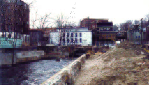 riverwalk (2) A river flows alongside a row of buildings and bare trees, with a dirt embankment in the foreground.