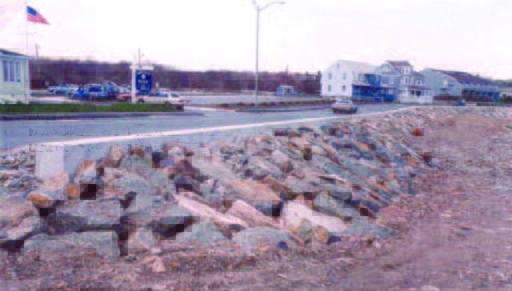 seawall (2) A rocky embankment lines a road near buildings, with cars parked and an American flag visible in the background.